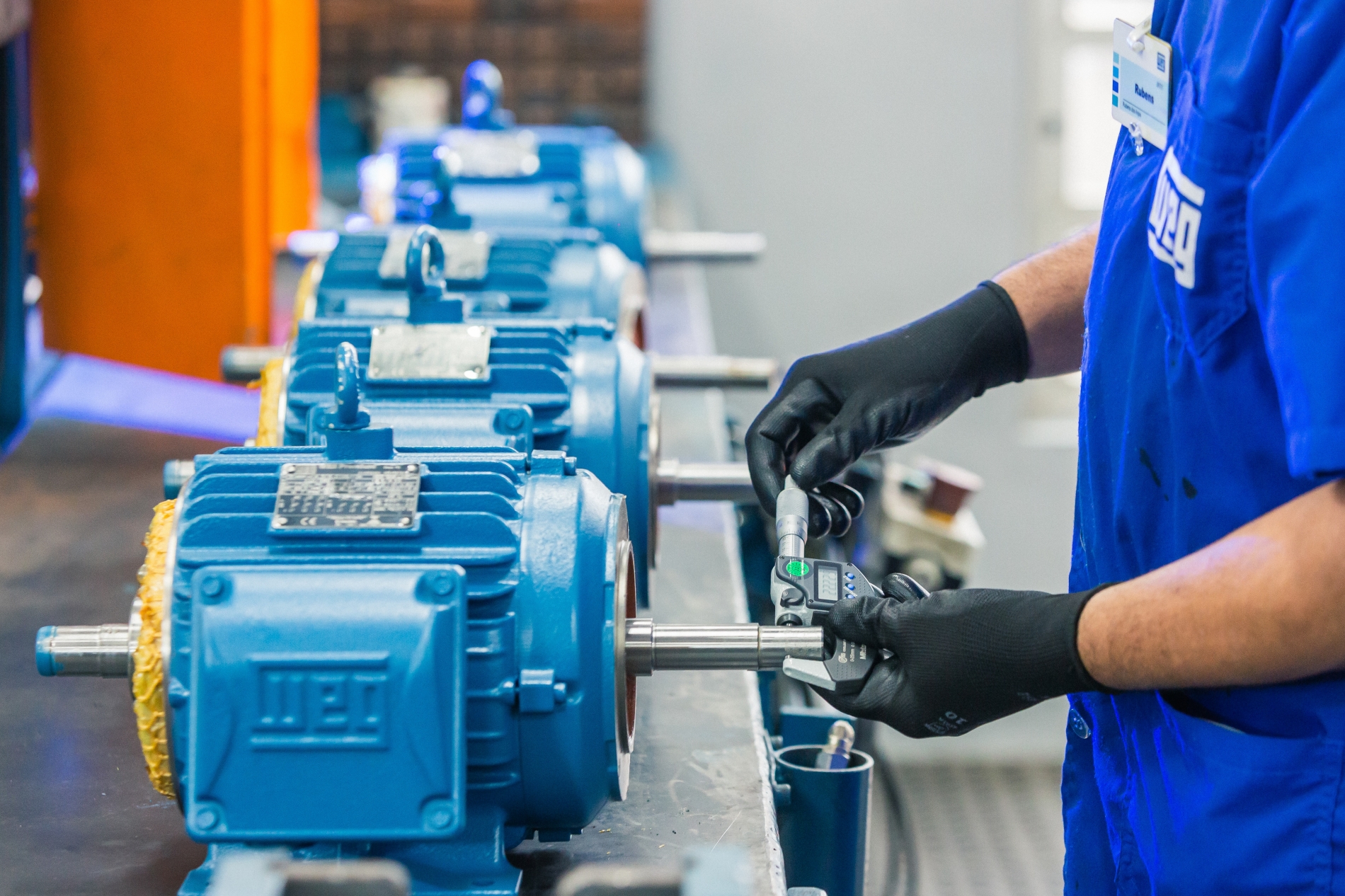 Technician performing precision shaft measurements on a series of blue WEG electric motors using a digital micrometer in a workshop.