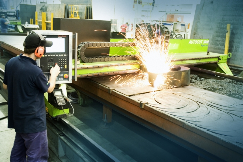 A worker operating an industrial automation system in a factory, overseeing robotic arms assembling products with sparks flying from a CNC machine.